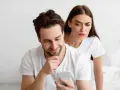 Une femme regarde par-dessus l'épaule de son partenaire qui utilise son téléphone, l'air suspicieuse. Elle essaie de calmer sa jalousie.