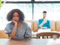Une jeune femme songeuse, le menton appuyé sur la main, semble réfléchir à un **problème de couple à qui en parler**. Son compagnon est flou en arrière-plan.
