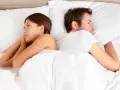Un couple allongé dos à dos dans un lit blanc, chacun perdu dans ses pensées, un soupir amoureux flotte dans l'air.