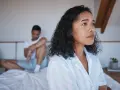 Une femme pensif sur le lit, son partenaire en arrière-plan. Elle pense : il est gentil mais je m'ennuie.