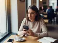Femme pensive au café, son téléphone à la main. Elle se demande comment savoir si un homme voit plusieurs femmes, cherchant des indices dans ses messages.