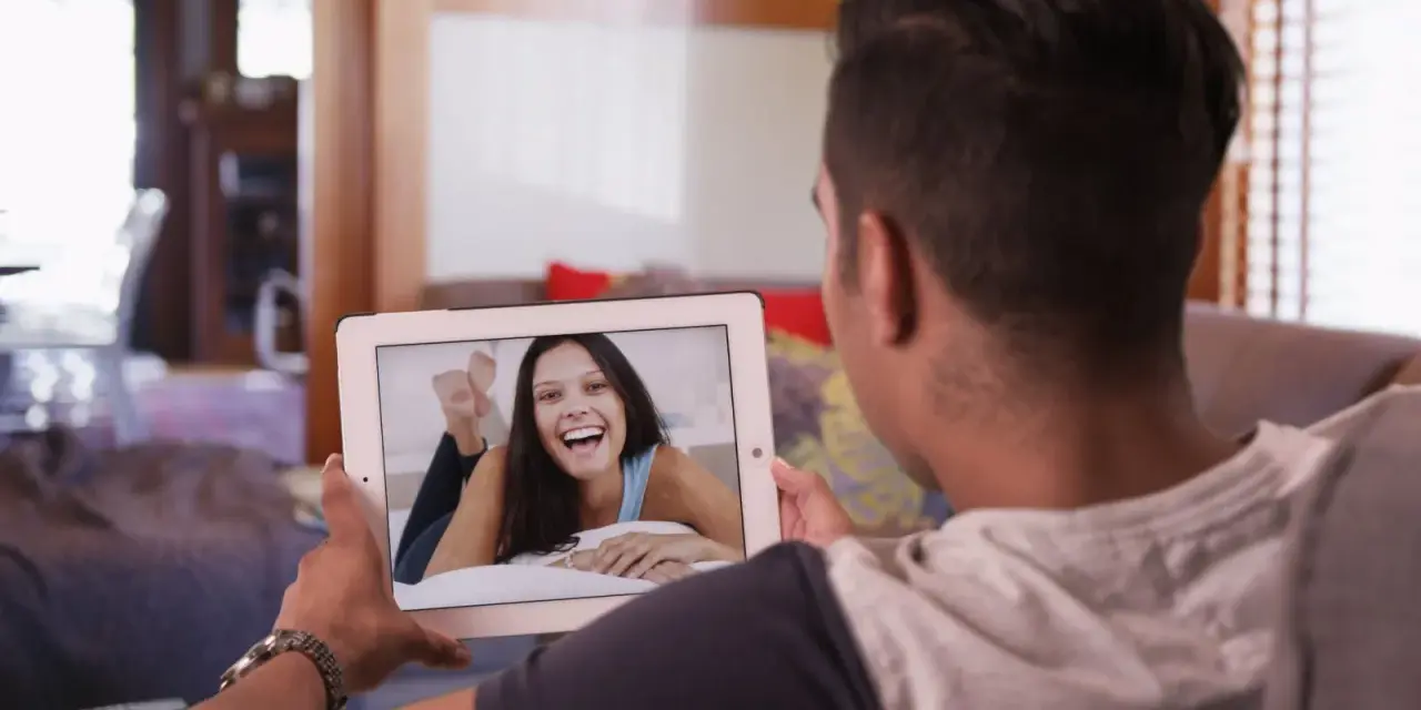 Un homme en relation à distance profite d'un appel vidéo avec sa compagne, dont le sourire illumine l'écran de sa tablette.