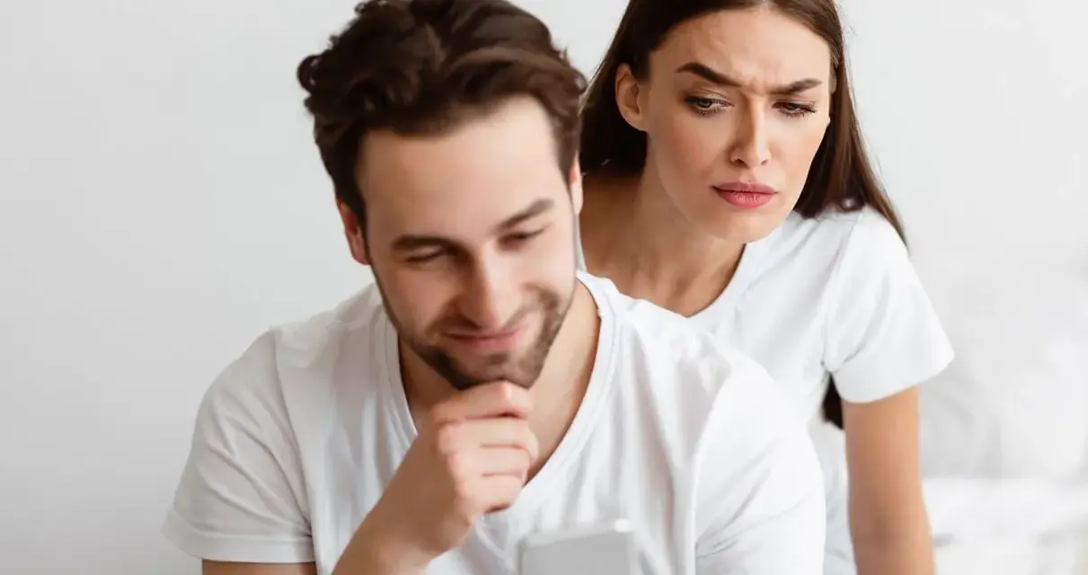 Une femme regarde par-dessus l'épaule de son partenaire qui utilise son téléphone, l'air suspicieuse. Elle essaie de calmer sa jalousie.