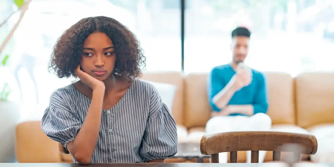 Une jeune femme songeuse, le menton appuyé sur la main, semble réfléchir à un **problème de couple à qui en parler**. Son compagnon est flou en arrière-plan.