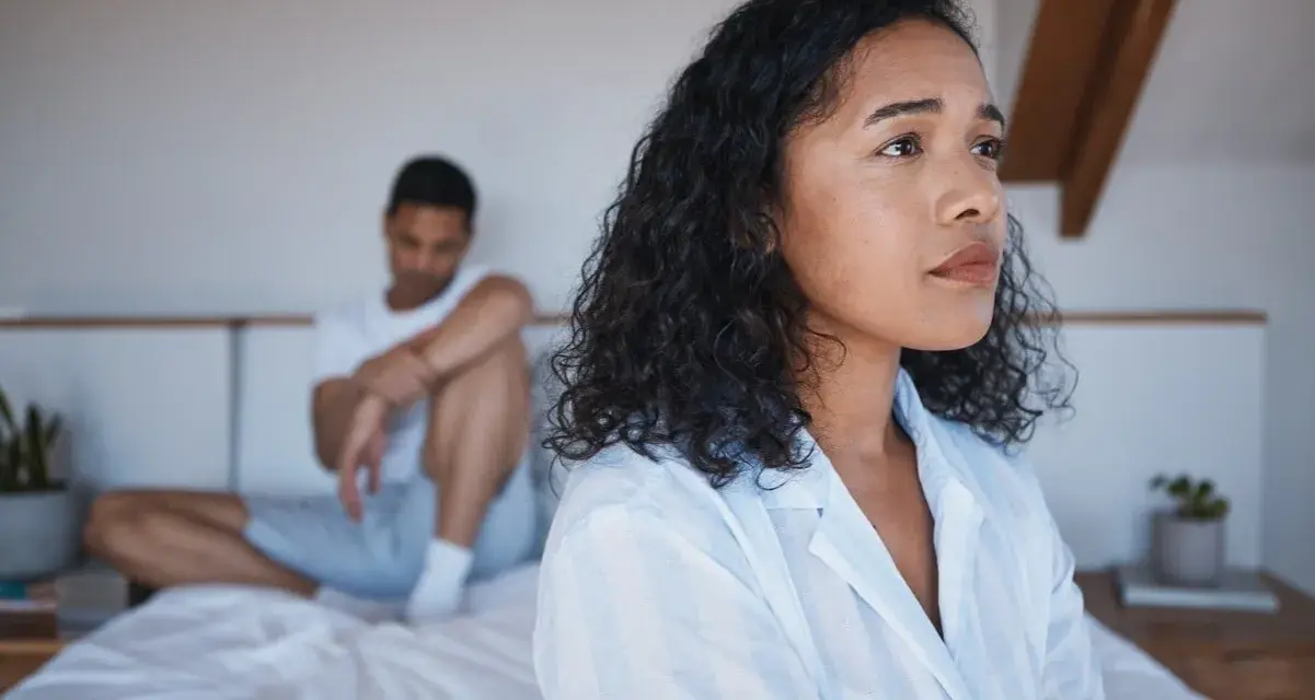 Une femme pensif sur le lit, son partenaire en arrière-plan. Elle pense : il est gentil mais je m'ennuie.