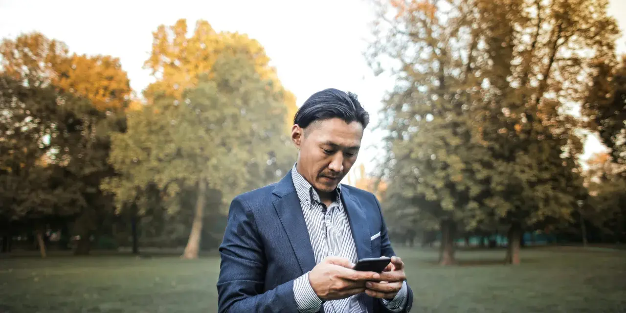 Un homme en costume, concentré sur son téléphone, dans un parc automnal. Une phrase provocante pour un homme pourrait être écrite sur son écran.