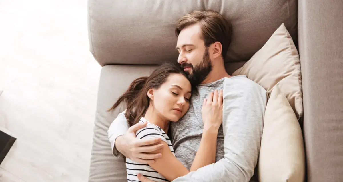Un couple dort paisiblement sur un canapé. L'homme enlace la femme, une scène qui peut évoquer une baisse de libido chez l'homme, mais aussi un profond sentiment de sécurité et d'amour partagé.