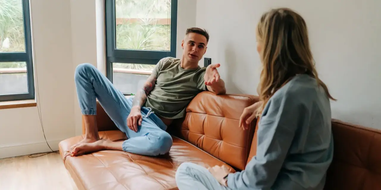 Un homme et une femme discutent sur un canapé en cuir marron. Leurs vêtements décontractés suggèrent un moment de détente, peut-être un double lien familial ou amical.