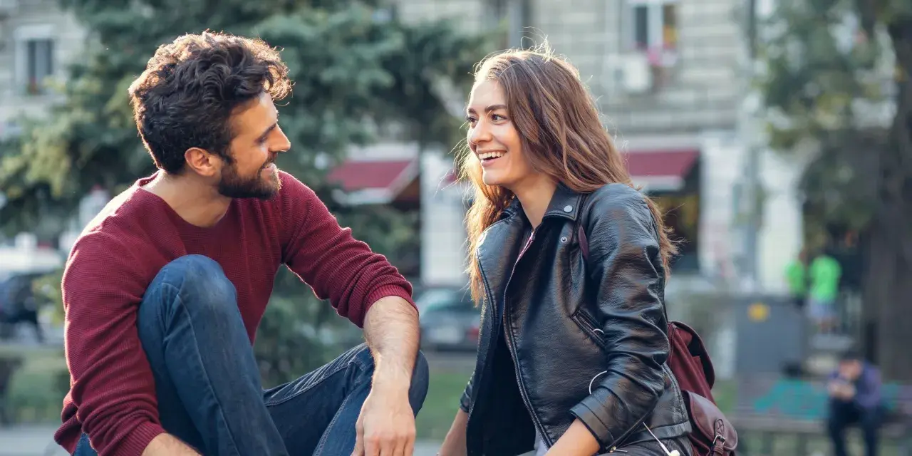 Un homme qui veut vous voir, assis sur l'herbe, rit avec une jeune femme. Ils semblent partager un moment complice et joyeux.