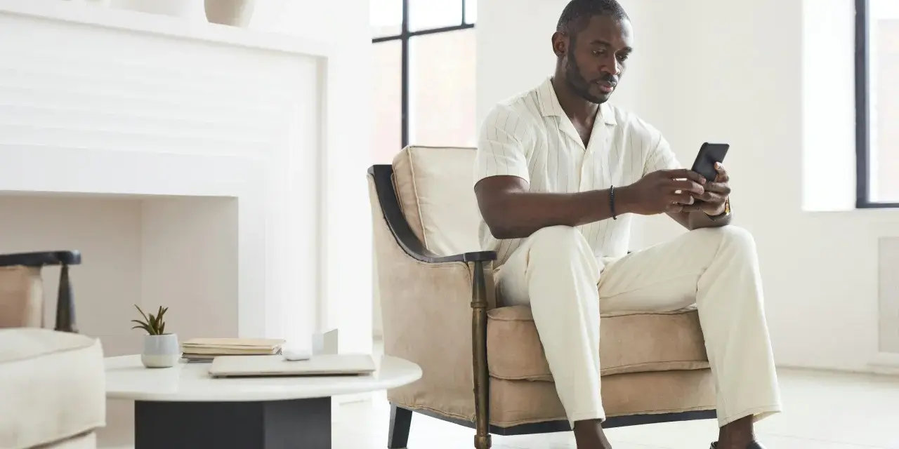 Un homme en tenue claire, assis dans un fauteuil, regarde son téléphone. Il attend un message, car il ne m'écrit plus comme avant.