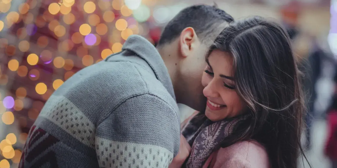 Un homme murmure à l'oreille d'une femme qui sourit, une scène où l'on peut être attiré par quelqu'un sans savoir pourquoi, sous des lumières festives.