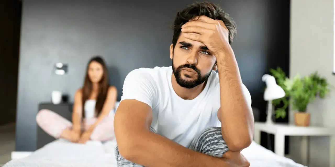 Un homme pensif sur un lit, sa compagne floue derrière. Comment attiser le désir d'un homme ? Peut-être par la communication et la connexion.