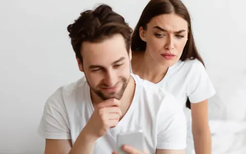Une femme regarde par-dessus l'épaule de son partenaire qui utilise son téléphone, l'air suspicieuse. Elle essaie de calmer sa jalousie.