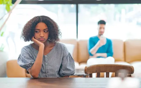 Une jeune femme songeuse, le menton appuyé sur la main, semble réfléchir à un **problème de couple à qui en parler**. Son compagnon est flou en arrière-plan.