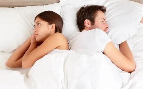 Un couple allongé dos à dos dans un lit blanc, chacun perdu dans ses pensées, un soupir amoureux flotte dans l'air.