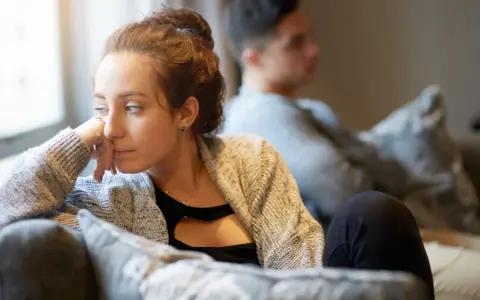 Une femme songeuse, le regard perdu, semble ressentir une profonde insécurité affective. Un homme est assis derrière elle, le dos tourné.