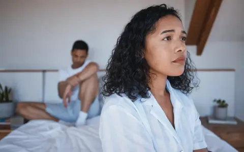 Une femme pensif sur le lit, son partenaire en arrière-plan. Elle pense : il est gentil mais je m'ennuie.