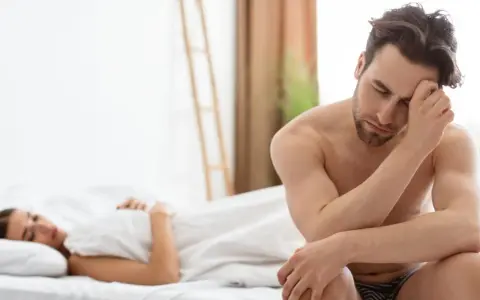 Un homme pensif, le regard baissé, semble préoccupé par un besoin sexuel non satisfait, tandis qu'une femme dort paisiblement à ses côtés.