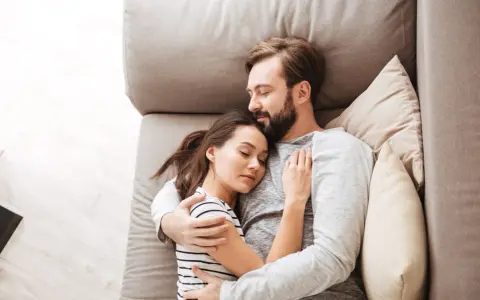 Un couple dort paisiblement sur un canapé. L'homme enlace la femme, une scène qui peut évoquer une baisse de libido chez l'homme, mais aussi un profond sentiment de sécurité et d'amour partagé.
