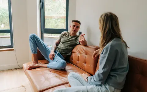 Un homme et une femme discutent sur un canapé en cuir marron. Leurs vêtements décontractés suggèrent un moment de détente, peut-être un double lien familial ou amical.