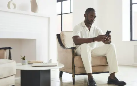 Un homme en tenue claire, assis dans un fauteuil, regarde son téléphone. Il attend un message, car il ne m'écrit plus comme avant.