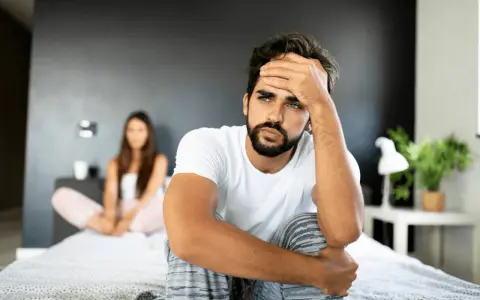 Un homme pensif sur un lit, sa compagne floue derrière. Comment attiser le désir d'un homme ? Peut-être par la communication et la connexion.