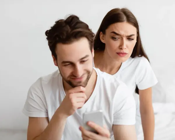 Une femme regarde par-dessus l'épaule de son partenaire qui utilise son téléphone, l'air suspicieuse. Elle essaie de calmer sa jalousie.