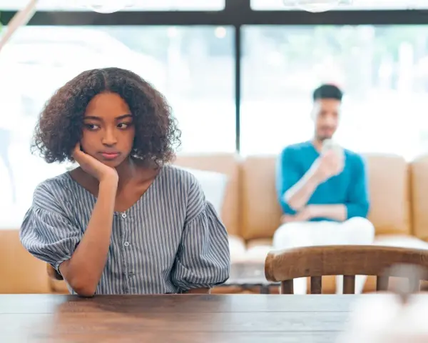 Une jeune femme songeuse, le menton appuyé sur la main, semble réfléchir à un **problème de couple à qui en parler**. Son compagnon est flou en arrière-plan.