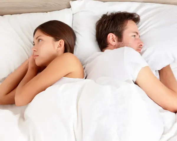 Un couple allongé dos à dos dans un lit blanc, chacun perdu dans ses pensées, un soupir amoureux flotte dans l'air.