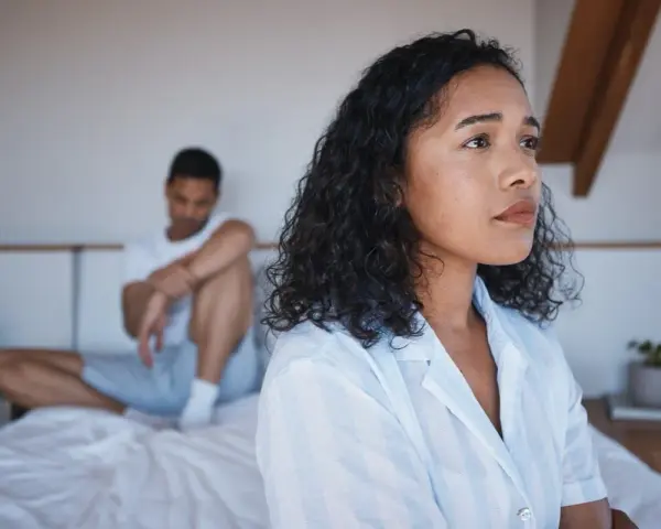 Une femme pensif sur le lit, son partenaire en arrière-plan. Elle pense : il est gentil mais je m'ennuie.