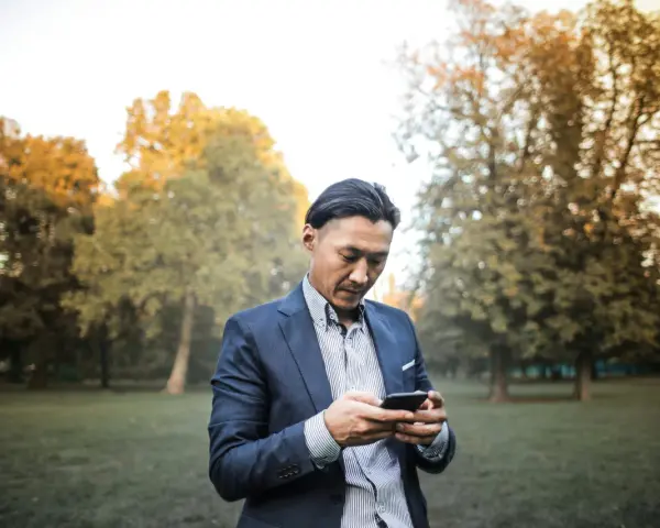 Un homme en costume, concentré sur son téléphone, dans un parc automnal. Une phrase provocante pour un homme pourrait être écrite sur son écran.
