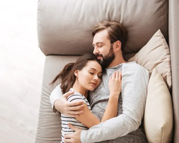 Un couple dort paisiblement sur un canapé. L'homme enlace la femme, une scène qui peut évoquer une baisse de libido chez l'homme, mais aussi un profond sentiment de sécurité et d'amour partagé.