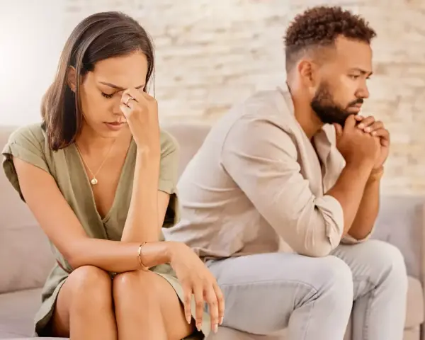 Un couple assis dos à dos, visiblement affecté par des disputes incessantes. La femme se tient la tête, l'air accablé, tandis que l'homme fixe le vide, les mains jointes.