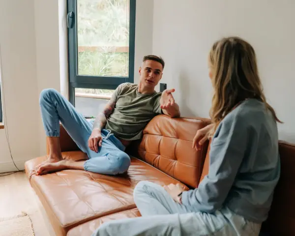 Un homme et une femme discutent sur un canapé en cuir marron. Leurs vêtements décontractés suggèrent un moment de détente, peut-être un double lien familial ou amical.