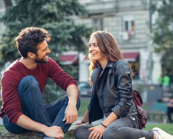 Un homme qui veut vous voir, assis sur l'herbe, rit avec une jeune femme. Ils semblent partager un moment complice et joyeux.