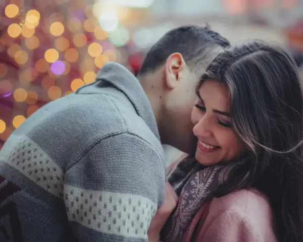 Un homme murmure à l'oreille d'une femme qui sourit, une scène où l'on peut être attiré par quelqu'un sans savoir pourquoi, sous des lumières festives.