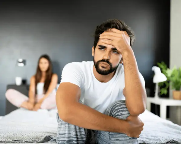 Un homme pensif sur un lit, sa compagne floue derrière. Comment attiser le désir d'un homme ? Peut-être par la communication et la connexion.