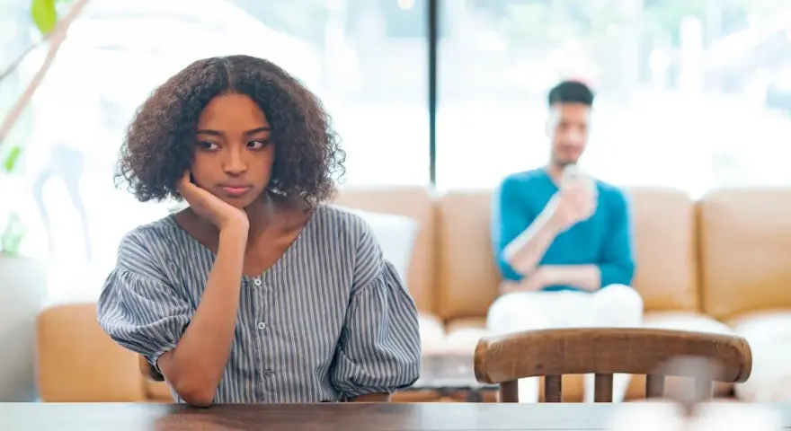 Une jeune femme songeuse, le menton appuyé sur la main, semble réfléchir à un **problème de couple à qui en parler**. Son compagnon est flou en arrière-plan.