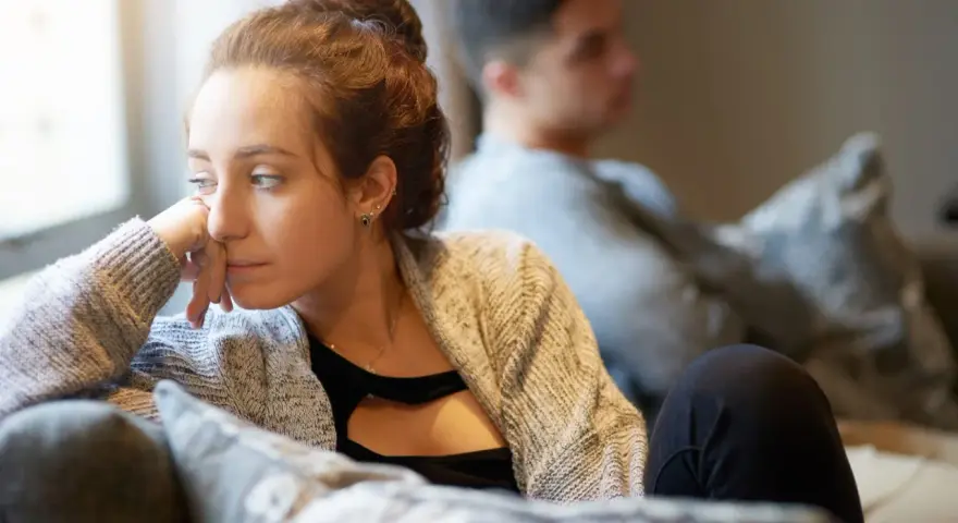 Une femme songeuse, le regard perdu, semble ressentir une profonde insécurité affective. Un homme est assis derrière elle, le dos tourné.