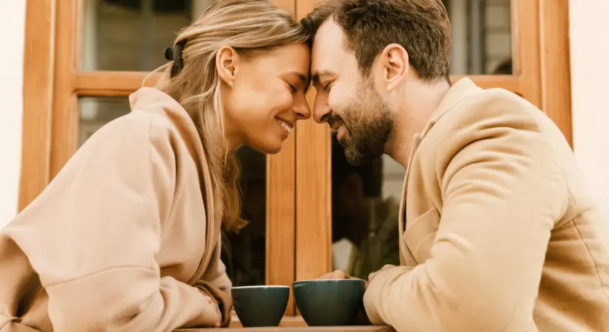 Un couple partage un moment intime, leurs fronts se touchant, illustrant l'effet miroir en amour. Deux tasses de café attendent sur la table.