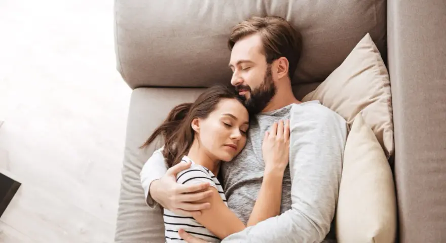 Un couple dort paisiblement sur un canapé. L'homme enlace la femme, une scène qui peut évoquer une baisse de libido chez l'homme, mais aussi un profond sentiment de sécurité et d'amour partagé.