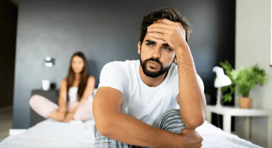Un homme pensif sur un lit, sa compagne floue derrière. Comment attiser le désir d'un homme ? Peut-être par la communication et la connexion.