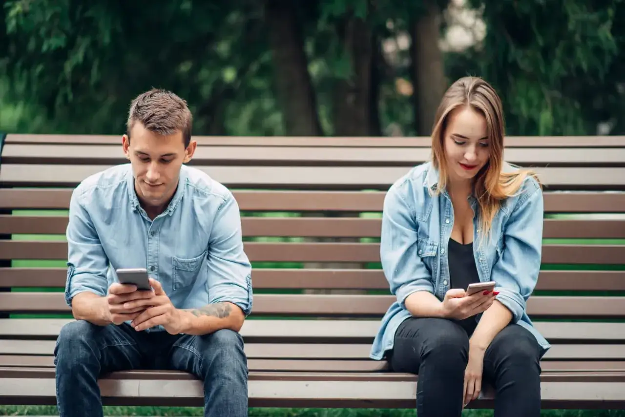 Un couple assis sur un banc, chacun absorb&eacute; par son t&eacute;l&eacute;phone. Un exemple de l'effet boomerang en amour, o&ugrave; la distance num&eacute;rique cr&eacute;e une distance r&eacute;elle.