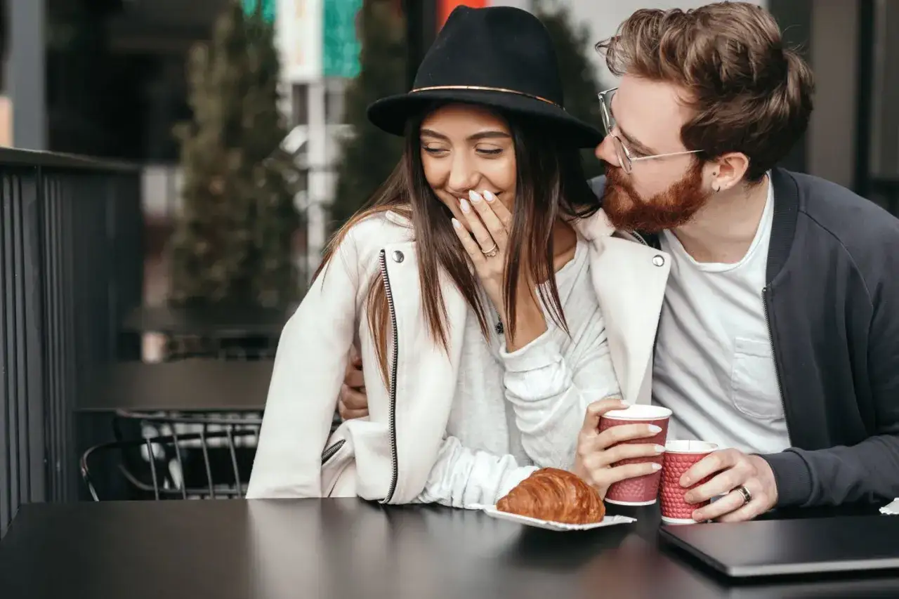 Un homme chuchote &agrave; l'oreille d'une femme qui rit, la main devant la bouche. Ils sont assis &agrave; une table avec des caf&eacute;s et un croissant, semblant faire attendre un homme.