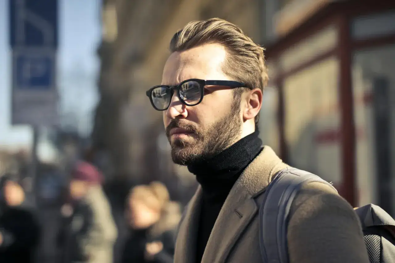 Un homme au regard insistant, portant des lunettes noires et un manteau beige, attend dans la rue.