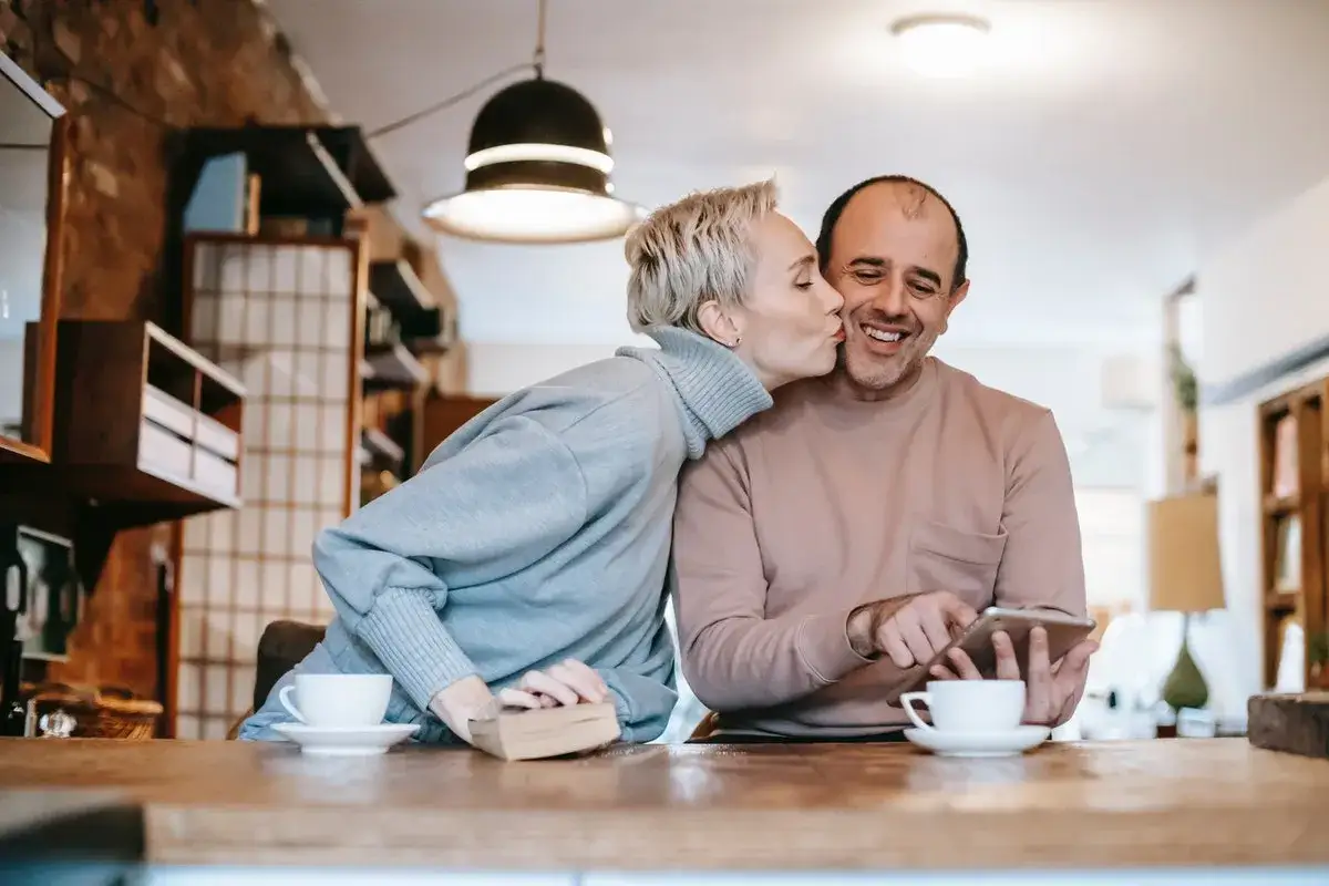 Un couple partage un moment tendre. Elle l'embrasse sur la joue, un livre et une tasse de café à proximité. Ils semblent apprendre à se connaître, profitant de leur temps ensemble.