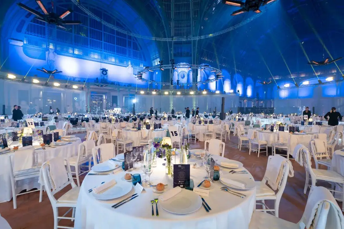 Salle de r&eacute;ception &eacute;l&eacute;gante, tables dress&eacute;es pour un d&icirc;ner de gala. L'ambiance bleue sugg&egrave;re une soir&eacute;e o&ugrave; l'on pourrait rencontrer un homme riche.