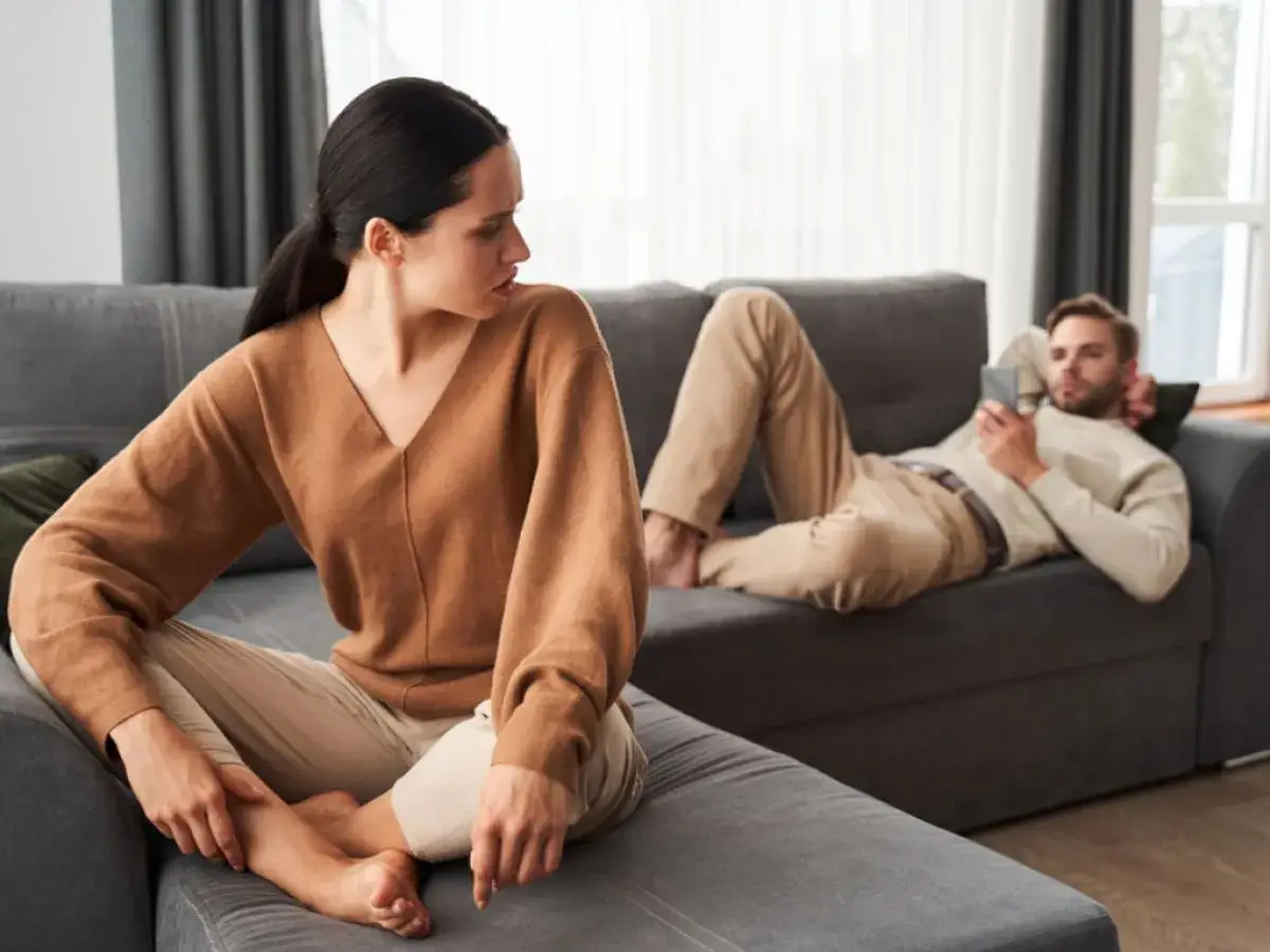 Une femme regarde son partenaire, qui est allong&eacute; sur le canap&eacute;, absorb&eacute; par son t&eacute;l&eacute;phone. La sc&egrave;ne illustre la psychologie du refus de communiquer dans un couple.