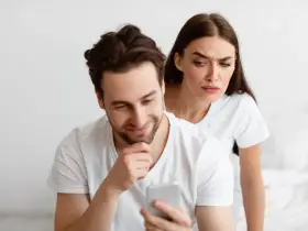 Une femme regarde par-dessus l'épaule de son partenaire qui utilise son téléphone, l'air suspicieuse. Elle essaie de calmer sa jalousie.
