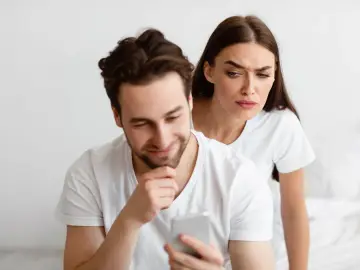 Une femme regarde par-dessus l'épaule de son partenaire qui utilise son téléphone, l'air suspicieuse. Elle essaie de calmer sa jalousie.